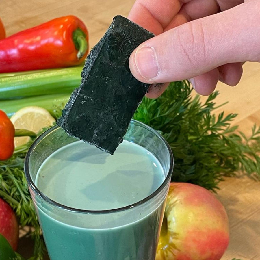One frozen Oasia Farms spirulina pod single serving held above a green smoothie with health foods behind depicting one way to eat spirulina
